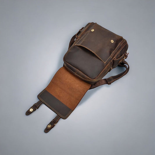 A top-down shot of a vintage-inspired leather backpack resting on a wooden surface. The bag's aged leather exterior displays a weathered patina, giving it a rustic and well-loved appearance. Distressed brass buckles and snap closures accentuate its antique charm, while the spacious interior and exterior pockets hint at its utility for travel or outdoor adventures.